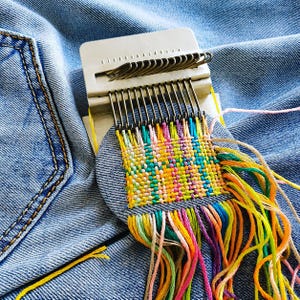 May include: A small plastic darning loom with metal hooks attached to blue denim fabric. The loom is actively weaving a square patch using multicoloured embroidery threads in shades of yellow, pink, green, and orange, with loose thread ends hanging below.