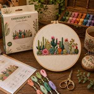May include: An embroidery kit featuring a cactus garden design. The set includes a cardboard box labelled EMBROIDERY KIT Cactus Garden, a circular wooden hoop with a finished cactus embroidery, a printed instruction sheet, colourful embroidery threads, and gold scissors.