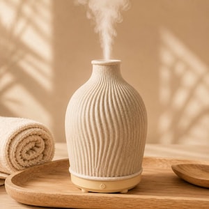 May include: A beige ceramic essential oil diffuser with a vertical wavy ribbed texture and a speckled finish. The diffuser has a narrow neck and a smooth base with a power button, emitting a steady stream of white mist from the top opening.