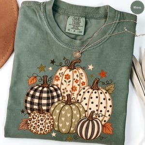 May include: A moss green t-shirt featuring a graphic print of five pumpkins with various patterns including black and white buffalo plaid, leopard print, polka dots, hearts, and floral designs, accented by small stars and autumn leaves.