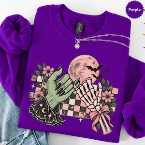 May include: A purple crewneck sweatshirt featuring a graphic print of a green hand with long black nails and a skeleton hand reaching toward each other. The design includes a pink moon, bats, flowers, and a black and white checkered pattern.
