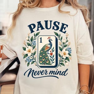 May include: An off-white t-shirt featuring a graphic print with the text PAUSE at the top and Never mind at the bottom. The center shows a rectangular tile with the number 1 and a peacock illustration, surrounded by floral branches with white flowers.