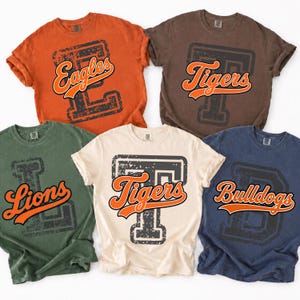 May include: Five short-sleeve crew neck t-shirts in orange, brown, olive green, cream, and navy blue. Each shirt features a large, distressed block letter background with a cursive team name graphic in orange and white, reading Eagles, Tigers, Lions, or Bulldogs.