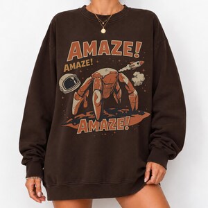 May include: A dark brown crewneck sweatshirt featuring a graphic print of a robotic creature on a planet surface, an astronaut helmet, and a small rocket. The text AMAZE! appears in large orange letters above and below the central illustration.