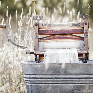 May include: An antique wooden washing machine with a metal tub, a crank handle, and a white cloth inside. The machine is labeled "Anchor Brand" and is sitting in a field of tall grass. The image is 8 inches by 10 inches.
