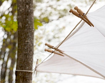 May include: A white fabric shelter is secured to a tree with wooden clothespins and rope. The scene is set outdoors with a blurred background of trees and foliage, suggesting a natural setting.