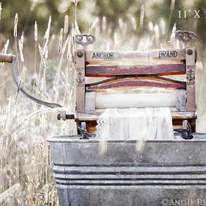 May include: An antique, wooden, hand-crank washing machine with a white cloth inside. The machine is labeled "Anchor Brand" and is sitting in a metal tub in a field of tall grass.
