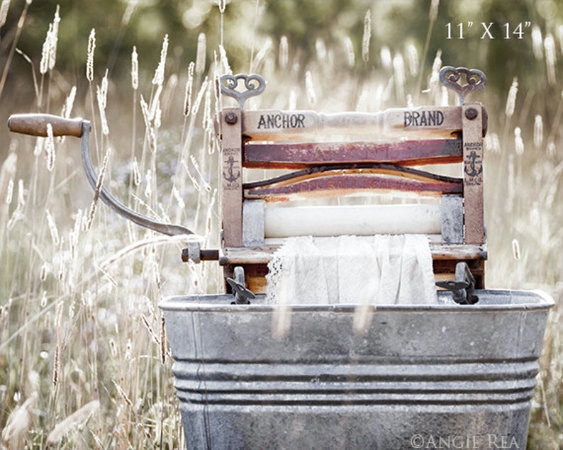 May include: An antique, wooden, hand-crank washing machine with a white cloth inside. The machine is labeled "Anchor Brand" and is sitting in a metal tub in a field of tall grass.