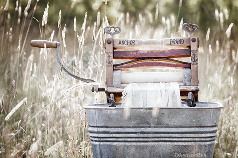 May include: An antique wooden washing machine with a metal tub and a crank handle. The machine is labeled "Anchor Brand" and has a white cloth inside. The machine is sitting in a field of tall grass.