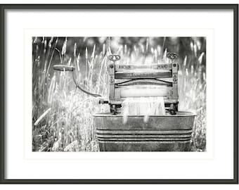 May include: Black and white photo of an antique Anchor Brand washing machine in a field of tall grass. The machine is in a metal tub and has a wooden handle.