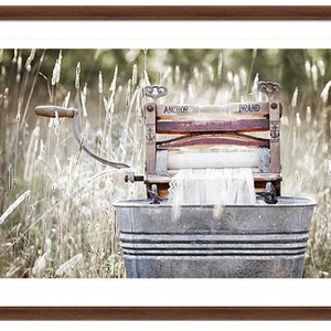 May include: An antique wooden washing machine with a metal tub, labeled "Anchor Brand", sits in a field of tall grass. The machine has a wooden roller and a crank handle.