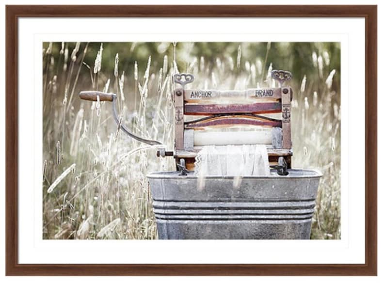 May include: An antique wooden washing machine with a metal tub, labeled "Anchor Brand", sits in a field of tall grass. The machine has a wooden roller and a crank handle.