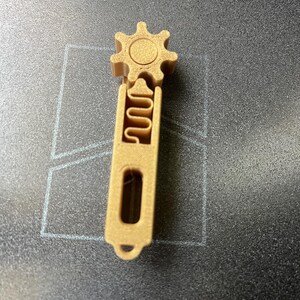 May include: A tan 3D-printed gear-shaped object with a handle. The gear has a circular center and six points. The handle has a rectangular slot and a loop at the bottom. The object is on a dark surface.
