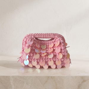 May include: A pink crocheted handbag featuring a thick braided handle and a structured rectangular body. The exterior is embellished with numerous round, iridescent, light-reflecting sequins that create a shimmering effect across the surface.