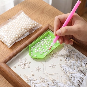 May include: A craft project in progress, featuring a framed artwork with a winter theme. The artwork is being embellished with white pearls, using a pink stylus and a green tray. A bag of pearls is also visible.