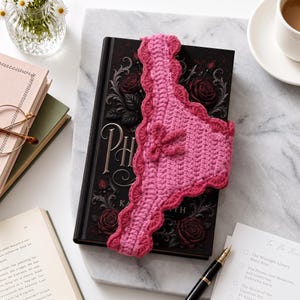 May include: A pink crocheted bookmark featuring a scalloped edge and a small bow detail, resting on top of a black hardcover book with a rose illustration. The bookmark is made of light and dark pink yarn with a textured, hand-stitched appearance.