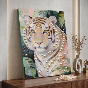 Puede incluir: Impresión artística en lienzo que presenta un tigre con adornos de perlas. El tigre tiene un pelaje blanco y canela con rayas negras y ojos amarillos. El fondo incluye follaje verde y tonos rosados suaves. La obra de arte se exhibe sobre una superficie de madera.
