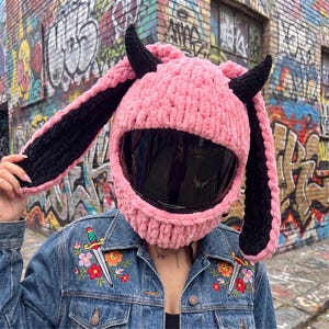 May include: A pink crochet helmet cover featuring long, black-lined bunny ears and two black horns. The cover is designed to fit over a full-face motorcycle helmet with a dark, reflective visor, creating a textured, plush appearance.