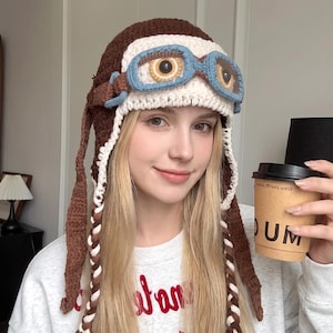 May include: A brown and cream crocheted aviator hat featuring integrated blue crocheted goggles with brown and yellow circular eye details. The hat includes long, braided side flaps with brown and cream yarn, extending down from the main cap structure.