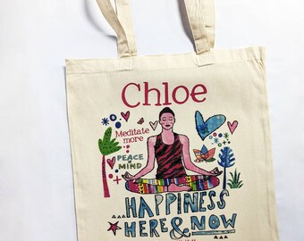 May include: A cream-colored canvas tote bag featuring a colorful illustration of a person meditating in a lotus position. The text reads Chloe, Meditate more, PEACE OF MIND, and HAPPINESS HERE & NOW, along with various small botanical and heart icons.