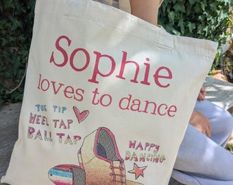 以下が含まれることがあります： 「Sophie loves to dance」というピンクの文字とタップシューズのイラストが描かれたクリーム色のキャンバストートバッグ。他にも「TOE TIP」「HEEL TAP」「BALL TAP」「HAPPY DANCING」という文字と、小さなピンクのハートと星がデザインされています。