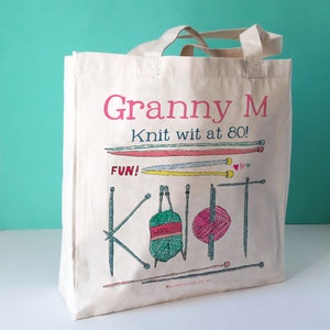 May include: A cream-coloured tote bag with a knitting-themed design. The bag displays the text "Granny M Knit wit at 80! FUN! Knit" in pink and green, alongside illustrations of knitting needles and yarn balls. It has a light beige handle.