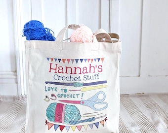 May include: A cream-colored canvas tote bag featuring a colorful graphic print with the text "Hannah's Crochet Stuff" and "LOVE TO CROCHET!". The design includes illustrations of crochet hooks, scissors, yarn, and bunting flags. Blue and pink yarn balls sit inside.