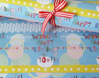 Puede incluir: Un rollo de papel de regalo azul claro con un patrón repetitivo del texto Whoopee Happy Birthday To You, círculos coloridos y banderines. Una cinta amarilla con estrellas blancas y un lazo a rayas rojas y blancas están atados alrededor.
