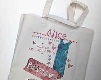 May include: A cream-colored canvas tote bag featuring the name Alice in pink text. The design includes a blue water bottle with the text DRINK ME!, a pink rolled-up mat labeled Pilates, and various small pink and blue stars and hearts.