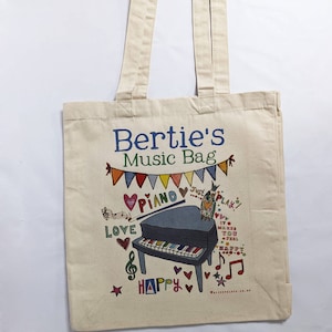 Puede incluir: Una bolsa de lona blanca con el texto "Bertie's Music Bag" impreso en azul. La bolsa presenta una ilustración colorida de un piano, notas musicales, corazones y las palabras "Love", "Piano", "Just Play", "It makes you feel happy" y "Happy".