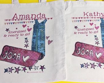 May include: Two white canvas tote bags featuring a printed design with the names Amanda and Kathy. The graphic includes a pink yoga mat, a blue water bottle with the text DRINK ME!, stars, hearts, bunting, and the text Energised & ready to go!