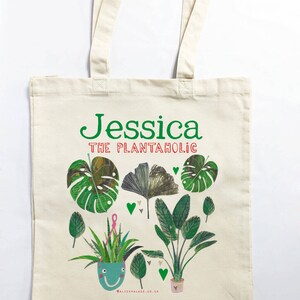 May include: A white canvas tote bag with green text that reads "Jessica The Plantaholic". The bag features a colorful illustration of various houseplants in pots with green leaves and pink hearts.