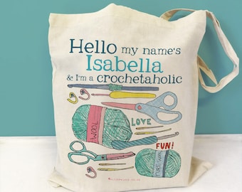 May include: A beige tote bag with the text "Hello my name's Isabella & I'm a crochetaholic" printed on it. The bag features a colorful illustration of yarn, scissors, crochet hooks, and needles.
