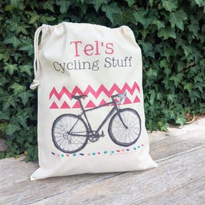 Puede incluir: Una bolsa de color crema con cordón que presenta el texto "Tel's Cycling Stuff" en rosa y gris. Debajo del texto hay un patrón de chevrón rosa y una ilustración negra de una bicicleta de carretera. El borde inferior tiene puntos coloridos.