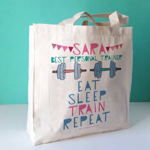 Op de afbeelding: Een witte canvas tote bag met roze en blauwe tekst die "Sara Best Personal Trainer Eat Sleep Train Repeat" zegt met een afbeelding van twee halters.