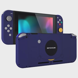 May include: A handheld gaming console in deep purple with a black screen, featuring a yellow analog stick, grey buttons, and a rear casing labeled SWITCHCUBE. The device includes a directional pad, four colored action buttons, and shoulder triggers.