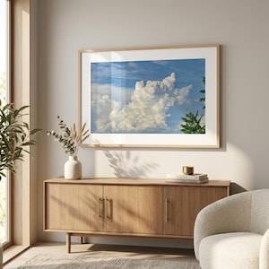 May include: A framed landscape wall art print featuring a large white cumulus cloud formation against a bright blue sky, with green tree foliage visible on the right side, displayed in a light wood frame with a white mat border.