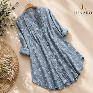 May include: A long-sleeved, button-down shirt in a dusty blue colour featuring a small white floral pattern. The shirt has a V-neckline with vertical pleats along the front placket and is displayed on a wooden hanger with the Lunaro Fashion logo.