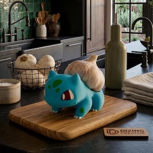 May include: A blue Bulbasaur-shaped kitchen gadget holding a bulb of garlic, resting on a wooden cutting board. The board has rounded edges and a small wooden sign that reads "DIZING CREATIVE". The kitchen setting includes a black sink, faucet, and other kitchen items.