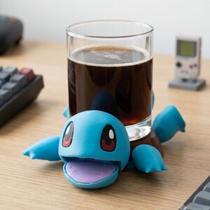 May include: A light blue turtle-shaped coaster with a smooth finish, large dark eyes, and an open mouth showing a purple interior. The coaster supports a clear glass filled with a dark brown liquid, featuring four flippers extending outward.