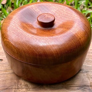 Pode incluir: Um recipiente redondo de madeira com tampa. O recipiente e a tampa são de cor castanha quente com um acabamento brilhante. A tampa tem um pequeno botão redondo na parte superior. O recipiente está sobre uma superfície de madeira.