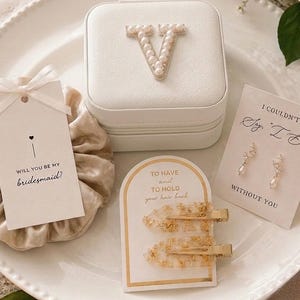 May include: A white square jewelry box with a pearl-encrusted letter V, a beige satin scrunchie with a tag reading Will you be my bridesmaid, two gold-toned hair clips on a card, and a pair of pearl earrings on a card reading I couldn't say I do without you.