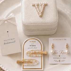 May include: A white square jewelry box with a pearl-encrusted letter V, two clear hair clips with gold flakes, and a pair of gold-tone pearl earrings. Includes three white cards with text: Will you be my bridesmaid, To have and to hold, and I couldn't tie the knot without you.