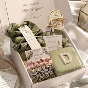 May include: A white gift box containing a sage green satin scrunchie, a set of four smaller satin scrunchies in pink, gray, and green, two gold-flecked hair clips, a pearl earring, and a sage green square jewelry case with a pearl-embellished letter D.