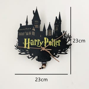 Puede incluir: Un reloj de pared negro con forma de castillo y el texto amarillo Harry Potter. El reloj tiene manecillas doradas finas y una pequeña figura colgante en la parte inferior. Las dimensiones indicadas son 23cm por 23cm.