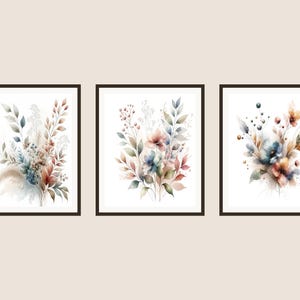 Puede incluir: Conjunto de tres láminas artísticas florales en acuarela enmarcadas. Cada lámina presenta una disposición única de flores y follaje en colores suaves y apagados, incluyendo azul, melocotón y marrón. La obra de arte se muestra sobre un fondo blanco.
