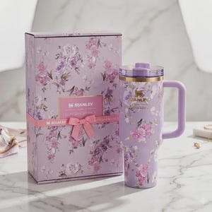 May include: A lavender Stanley travel mug with a handle and floral pattern, alongside a matching gift box. Both the mug and box display a repeating design of pink and purple flowers. The box is tied with a pink ribbon and has the text "STANLEY LOVESHACKFANCY".