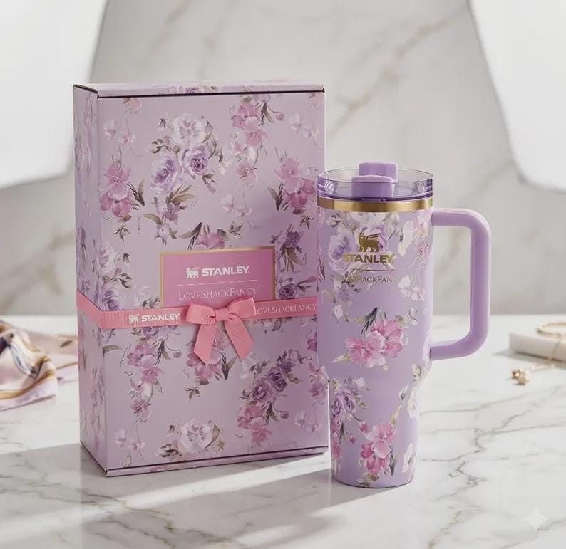 May include: A lavender Stanley travel mug with a handle and floral pattern, alongside a matching gift box. Both the mug and box display a repeating design of pink and purple flowers. The box is tied with a pink ribbon and has the text "STANLEY LOVESHACKFANCY".