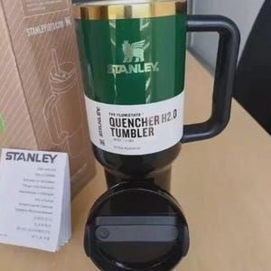 Puede incluir: Un vaso Stanley Quencher H2.0 verde y negro con asa y tapa negras. El vaso tiene un borde dorado y el logotipo de Stanley. El texto "THE FLOWSTATE QUENCHER H2.0 TUMBLER" est&aacute; impreso en el vaso.