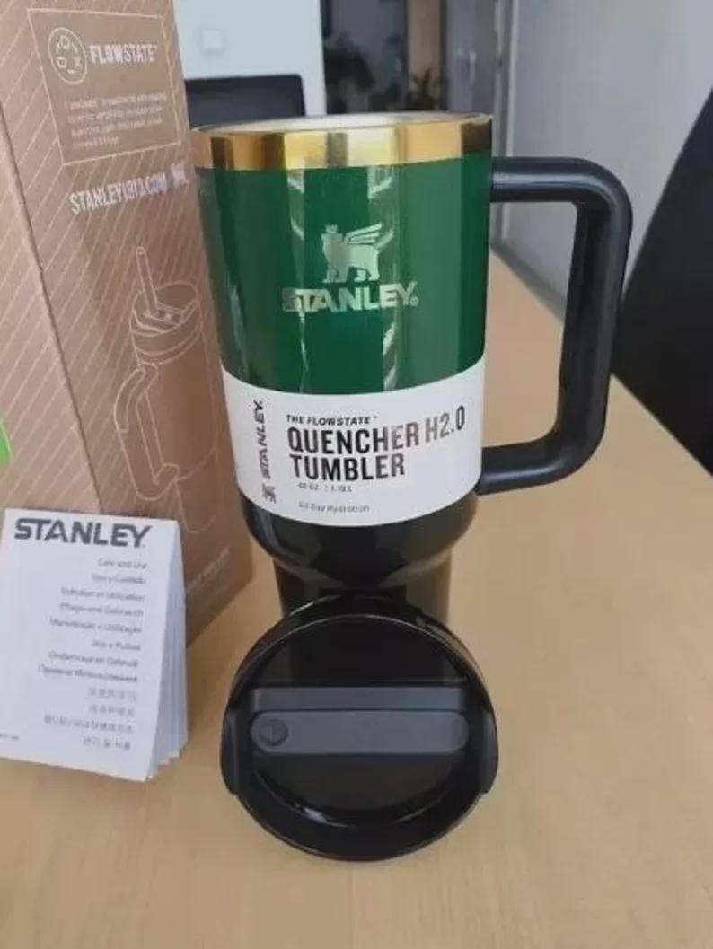 Puede incluir: Un vaso Stanley Quencher H2.0 verde y negro con asa y tapa negras. El vaso tiene un borde dorado y el logotipo de Stanley. El texto "THE FLOWSTATE QUENCHER H2.0 TUMBLER" est&aacute; impreso en el vaso.